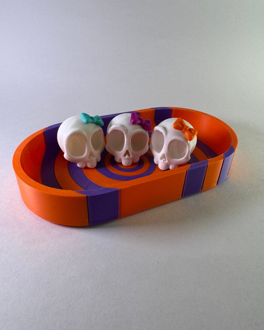 Tres calaveritas con moños de colores sobre una base naranja y morada.